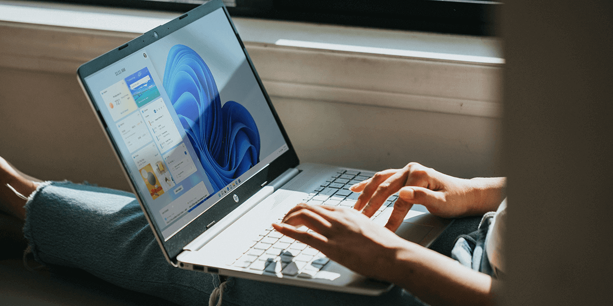 A laptop displays a Windows 11 interface; hands type on its keyboard; it rests on a person’s lap near a window